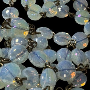 May include: A close-up of a vintage necklace featuring iridescent, light blue faceted beads. The beads are strung together with antique-looking metal links. The necklace is displayed against a black background, highlighting the beads' color and texture.