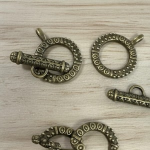 May include: Several antique bronze toggle clasps. The clasps feature a round ring with a decorative pattern and a cylindrical bar. The clasps are designed to connect jewelry pieces. The clasps are on a light brown wooden surface.