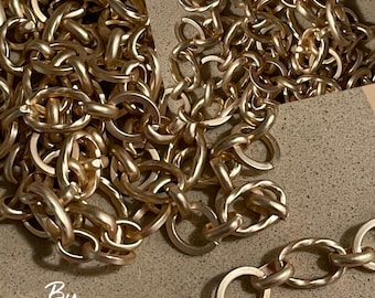 May include: Close-up of a gold-colored chain with oval and round links. The chain is arranged in a tangled pattern, with some sections overlapping. The text "By Pluffbeads" is visible in the lower left corner.