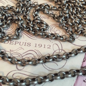 Pode incluir: Um colar de corrente de prata com um design simples de elos redondos. A corrente est&aacute; sobre um peda&ccedil;o de papel com o texto "DEPUIS 1912" impresso nele.