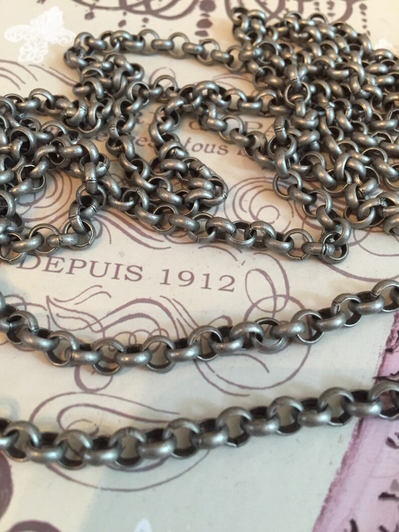 Pode incluir: Um colar de corrente de prata com um design simples de elos redondos. A corrente est&aacute; sobre um peda&ccedil;o de papel com o texto "DEPUIS 1912" impresso nele.