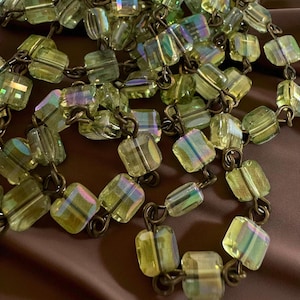 May include: A necklace featuring iridescent, square-cut beads in shades of green and yellow. The beads are strung together with dark metal links, creating a vintage or antique aesthetic. The beads have a subtle, shimmering quality.