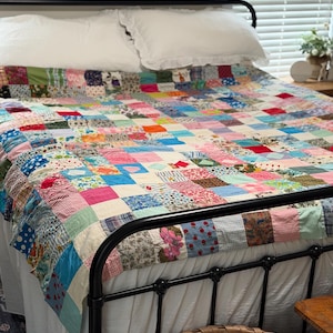 May include: A patchwork quilt featuring a grid of numerous small fabric squares in a wide variety of colors, patterns, and floral prints. The quilt is draped over a bed with white pillows and a black metal frame, displaying a multi-colored design.
