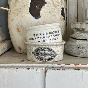 May include: Two vintage ceramic jars with white glaze and black lettering. The top jar reads "Banks & Cooke's Home Potted Beef Shrimps West Kirby". The bottom jar reads "W. Plumtree Ltd. Manufacturer of Home Potted Meats Railway St. Southport".