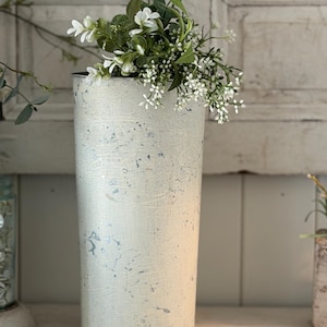 May include: A tall, cylindrical vase with a distressed, off-white finish and subtle blue accents. The vase is filled with a bouquet of white and green artificial flowers, creating a rustic, farmhouse aesthetic. Ideal for home decor.