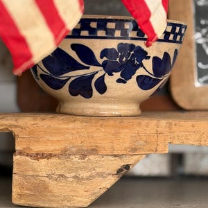 May include: A beige ceramic bowl featuring a dark blue floral pattern around the center and a checkered border near the rim. The bowl has a rounded base and sits on a wooden surface, partially obscured by a red and white striped fabric element.