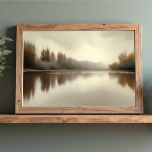 Puede incluir: Una impresión artística enmarcada que presenta una serena escena lacustre con árboles y un cielo nublado. La obra de arte se exhibe en una estantería de madera, flanqueada por plantas en macetas en jarrones blancos. El marco es de madera clara.