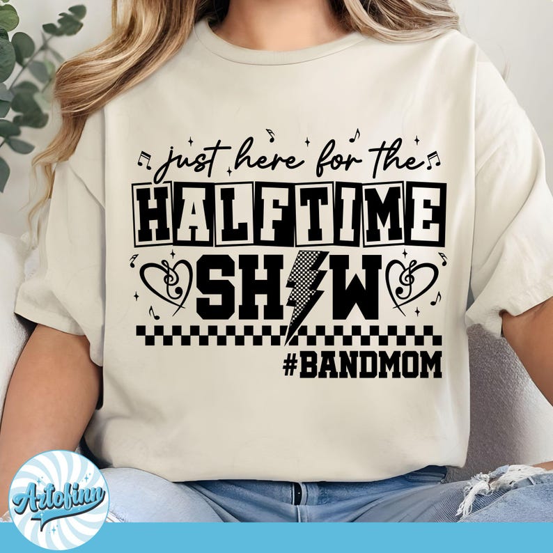 May include: Cream-colored t-shirt with the text "just here for the HALFTIME SHOW" in a black, bold font. The design includes musical notes, hearts, and a lightning bolt. The hashtag #BANDMOM is at the bottom.