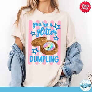 May include: Cream-colored t-shirt with a graphic of a dumpling in a bamboo steamer. The text reads "you're a glitter DUMPLING" in blue, with pink stripes and blue stars. The design is a PNG design.