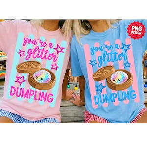 Puede incluir: Dos camisetas, una rosa y otra azul, cada una con un gráfico de una empanadilla en un vaporizador de bambú. El texto "you're a glitter DUMPLING" está encima y debajo de la ilustración de la empanadilla. El diseño incluye estrellas y rayas verticales.