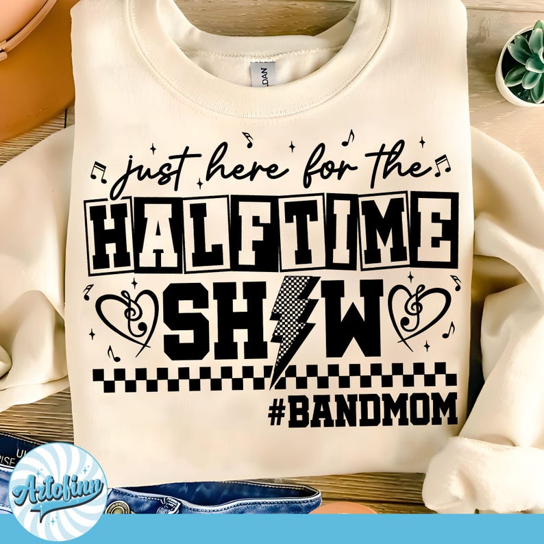 May include: Cream-colored sweatshirt with a black graphic that reads "just here for the HALFTIME SHOW" with musical notes and a lightning bolt. The text "#BANDMOM" is below the main text. A casual, comfortable top.