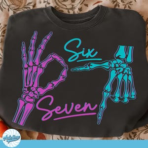 May include: Dark gray sweatshirt with a graphic design featuring skeleton hands forming the numbers six and seven. The word "Six" is in teal script, and "Seven" is in pink script. The skeleton hands are in pink and teal.