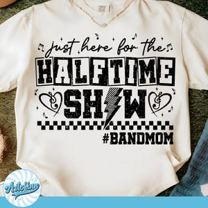 May include: Cream-colored t-shirt with a black graphic that reads "just here for the HALFTIME SHOW" with musical notes and a lightning bolt. The text "#BANDMOM" is below. A gold necklace is visible at the neckline.