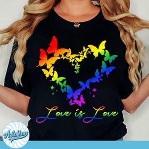 Puede incluir: Camiseta negra con un diseño en forma de corazón hecho de mariposas coloridas en tonos arcoíris. Las palabras "Love is Love" están escritas en una fuente degradada de arcoíris debajo.