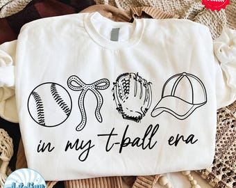 May include: White sweatshirt with a baseball, bow, glove, and cap graphic. The text "in my t-ball era" is printed below the graphics in a handwritten style. The design is in black.