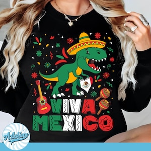 Puede incluir: Camiseta negra de manga larga con un gráfico colorido que presenta un dinosaurio verde con sombrero, una guitarra, maracas y el texto "VIVA MEXICO". El dinosaurio sostiene un corazón con la bandera mexicana.