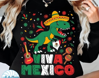 Pode incluir: Camiseta preta de manga comprida com um gráfico colorido com um dinossauro verde usando um chapéu, uma guitarra, maracas e o texto "VIVA MEXICO". O dinossauro segura um coração com a bandeira mexicana.
