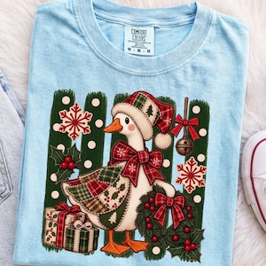 Könnte beinhalten: Ein hellblaues T-Shirt mit einer Grafik einer weißen Gans, die eine rot-grün karierte Weihnachtsmütze und eine passende Schleife trägt. Die Gans ist umgeben von roten Schneeflocken, einem grünen Kranz mit roter Schleife und zwei Geschenkpaketen.