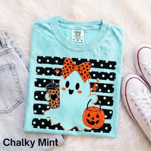 Könnte beinhalten: Ein hellblaues T-Shirt mit einer Grafik eines weißen Geistes, der eine orange-schwarze gepunktete Schleife trägt. Der Geist hält einen Kaffeebecher mit Leopardenmuster und einen orangefarbenen Kürbiseimer vor einem schwarz-weiß gepunkteten Hintergrund.
