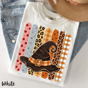 Könnte beinhalten: Ein weißes T-Shirt mit einem Grafikdruck eines dunkelbraunen Hexenhuts mit einem orange-weiß karierten Band. Der Hut ist über vertikalen Pinselstrich-Streifen mit Mustern aus roten Punkten, Leopardenmuster und orangefarbenem Schottenkaro platziert.