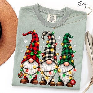 Può includere: Una t-shirt verde salvia con la grafica di tre gnomi che indossano cappelli a quadri rossi, neri e verdi. Gli gnomi hanno barbe bianche e sono avvolti da luci natalizie colorate con fiocchi di neve bianchi sullo sfondo. L'etichetta riporta Comfort Colors.