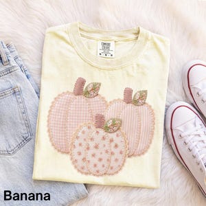 Könnte beinhalten: Ein blassgelbes T-Shirt mit einer Grafik von drei Kürbissen mit genähten Kanten. Die Kürbisse sind mit hellrosa Vichy-Karo-, Streifen- und Blumenmustern verziert. Das T-Shirt hat einen Rundhalsausschnitt und ein Etikett mit Comfort Colors.