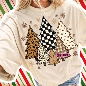 Könnte beinhalten: Ein cremefarbenes T-Shirt mit einer Grafik aus mehreren stilisierten Weihnachtsbäumen mit verschiedenen Mustern wie Leopardenmuster, schwarz-weißem Schachbrettmuster, Punkten und Karomuster, akzentuiert durch Schneeflocken und ein rosa Etikett mit der Aufschrift DON'T COPY.