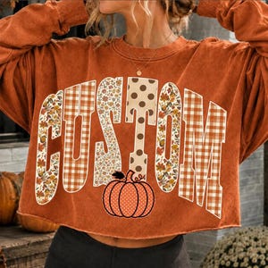 Könnte beinhalten: Ein langärmeliges Sweatshirt in gebranntem Orange mit dem Wort CUSTOM in großen Blockbuchstaben. Die Buchstaben haben verschiedene Muster, darunter Blumenprints, braun-weißes Vichy-Karo und braune Punkte. Darunter befindet sich eine Kürbisgrafik.