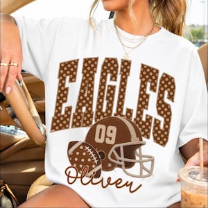 Könnte beinhalten: Ein weißes Kurzarm-T-Shirt mit dem Wort EAGLES in großen braunen gepunkteten Buchstaben. Unter dem Text befindet sich ein brauner Football-Helm mit der Nummer 09 und ein brauner gepunkteter Football, mit dem Namen Oliver in brauner Schreibschrift.