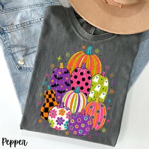 Könnte beinhalten: Ein anthrazitfarbenes T-Shirt mit einem bunten Grafikdruck, der mehrere Kürbisse in verschiedenen Farben und Mustern zeigt, darunter Punkte, Streifen, Blumen, Fledermäuse und Geister, akzentuiert durch kleine Sterne und Blüten.