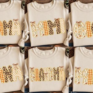 Könnte beinhalten: Eine Kollektion von sechs beigen Sweatshirts mit Rundhalsausschnitt, die jeweils ein anderes Wort in großen, gemusterten Blockbuchstaben zeigen. Die Wörter umfassen MAMA, MIMI, GIGI, NANA und GRANDMA. Jeder Buchstabe ist mit kleinen braunen Schleifen verziert.