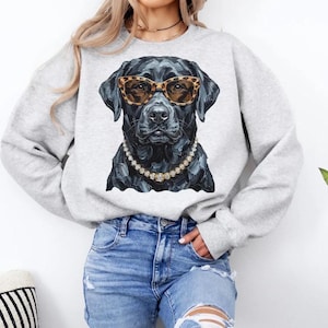 May include: A light gray crewneck sweatshirt featuring a graphic print of a black Labrador retriever wearing leopard print sunglasses and a beaded necklace. The dog is depicted in a detailed, painterly style with visible brushstrokes.