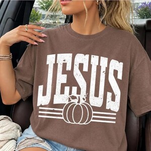 Könnte beinhalten: Ein braunes Kurzarm-T-Shirt mit dem Wort JESUS in großen, abgenutzten weißen Großbuchstaben. Unter dem Text befindet sich eine weiße Grafik eines Kürbisses, zentriert zwischen zwei Sätzen von drei horizontalen Linien in verwaschener Optik.