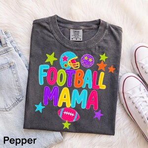 Könnte beinhalten: Ein anthrazitfarbenes Kurzarm-T-Shirt mit dem Schriftzug FOOTBALL MAMA in bunten Blockbuchstaben. Das Design zeigt einen Football-Helm, ein Smiley-Gesicht, einen Football und verschiedene Sterne in leuchtenden Neonfarben wie Pink, Blau, Gelb und Orange.