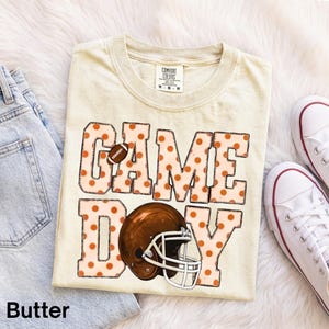 Könnte beinhalten: Ein cremefarbenes T-Shirt mit dem Schriftzug GAME DAY in großen Blockbuchstaben mit orangefarbenem Punktemuster. Der Buchstabe A ist durch einen Football ersetzt, und der Buchstabe O durch einen braunen Footballhelm mit weißem Gitter.
