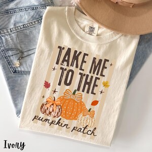 Könnte beinhalten: Ein elfenbeinfarbenes T-Shirt mit einem Aufdruck, der den Text "TAKE ME TO THE pumpkin patch" in dunkelbrauner Schrift zeigt. Das Design umfasst drei illustrierte Kürbisse in Orange und Weiß, akzentuiert durch kleine Herbstblätter und Sterne.