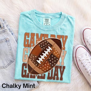 Könnte beinhalten: Ein hellblaues T-Shirt mit dem Schriftzug GAME DAY in braunen Blockbuchstaben. Mittig darüber befindet sich eine braune Football-Grafik mit weißen Punkten und weißen Schnürsenkeln. Das T-Shirt hat einen Rundhalsausschnitt und ein M-Größenetikett.