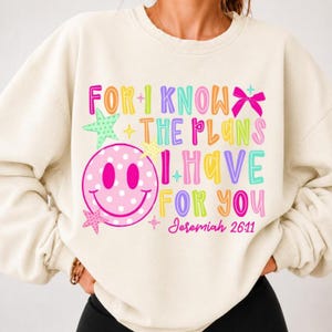 Könnte beinhalten: Ein cremefarbenes Sweatshirt mit Rundhalsausschnitt und einem bunten Grafikdruck mit dem Text "FOR I KNOW THE PLANS I HAVE FOR YOU Jeremiah 26:11". Das Design enthält ein großes rosa gepunktetes Smiley-Gesicht, grüne und rosa Sterne sowie eine rosa Schleife.