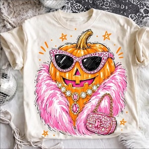 Könnte beinhalten: Ein cremefarbenes T-Shirt mit einer Grafik eines orangefarbenen Kürbisses, der eine pinke Glitzer-Katzenaugen-Sonnenbrille, eine pinke Kunstfell-Boa, eine Juwelenkette und eine passende pinke Glitzer-Handtasche trägt, umgeben von kleinen orangefarbenen Sternen.