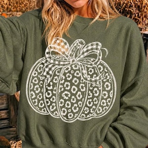 Könnte beinhalten: Ein olivgrünes Langarm-Sweatshirt mit einer großen weißen Grafik eines Kürbisses. Das Kürbisdesign zeigt ein Leopardenmuster auf dem Körper und ein Karomuster auf der Schleife, die oben am Kürbisstiel gebunden ist.