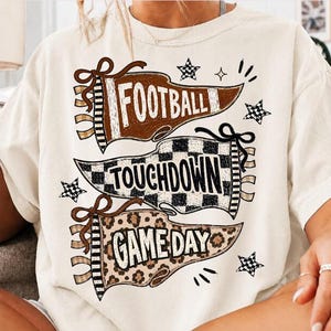 Könnte beinhalten: Ein cremefarbenes Kurzarm-T-Shirt mit einer Grafik aus drei Wimpeln. Der obere Wimpel ist braun mit dem Text FOOTBALL, der mittlere ist schwarz-weiß kariert mit TOUCHDOWN und der untere hat ein Leopardenmuster mit dem Text GAME DAY.
