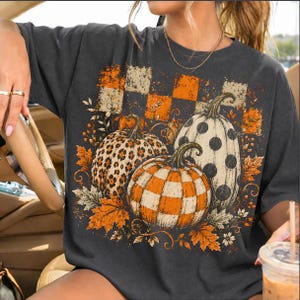 Könnte beinhalten: Ein anthrazitfarbenes Kurzarm-T-Shirt mit einem Grafikdruck von drei Kürbissen mit Leopardenmuster, Punkten sowie orange-weißem Karomuster, umgeben von Herbstblättern und einem karierten Hintergrunddesign.