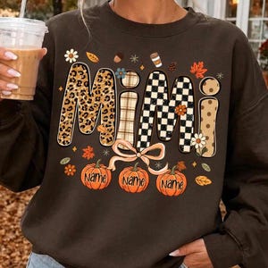 Könnte beinhalten: Ein dunkelbraunes Sweatshirt mit Rundhalsausschnitt und dem Wort MIMI in großen, gemusterten Buchstaben mit Leopardenmuster, Karomuster und Schachbrettmuster. Unter dem Text sind drei orangefarbene Kürbisse mit dem Wort NAME abgebildet.