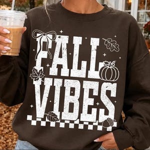 Könnte beinhalten: Ein dunkelbraunes Sweatshirt mit Rundhalsausschnitt und dem weißen, abgenutzten Schriftzug FALL VIBES. Die Grafik zeigt eine Schleife, einen Kürbis, Blätter, eine Eichel und kleine Sterne sowie ein Karomuster am unteren Rand.