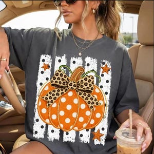 Könnte beinhalten: Ein anthrazitfarbenes Kurzarm-T-Shirt mit einer großen Grafik eines orangefarbenen Kürbisses mit weißen Punkten. Der Kürbis ist mit einer Schleife im Leopardenmuster verziert und vor vertikalen weißen Streifen mit schwarzen Punkten platziert.