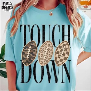 Könnte beinhalten: Ein hellblaues T-Shirt mit dem schwarzen Schriftzug TOUCH DOWN und drei Football-Grafiken. Die Footballs haben unterschiedliche Muster: eines mit Camouflage, eines mit Leopardenmuster und eines mit einem braun-beigen Karomuster.