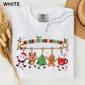 Könnte beinhalten: Ein weißes Kurzarm-T-Shirt mit einer großen goldenen Sicherheitsnadel auf der Brust. An der Nadel hängen fünf weihnachtliche Anhänger: ein Weihnachtsmann, ein Rentier, ein Tannenbaum, ein Lebkuchenmann und eine rote Tasse mit Kakao.