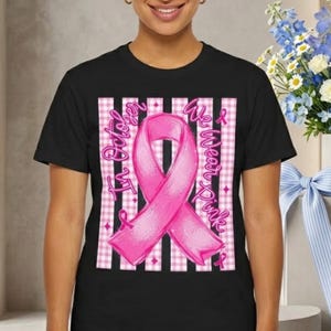 Op de afbeelding: Een zwart t-shirt met een grote roze bewustwordingslint gecentreerd op een wit en roze gingham gestreepte achtergrond. De tekst "In October We Wear Pink" staat in een cursief roze lettertype en buigt rond het lint op de voorkant van het shirt.