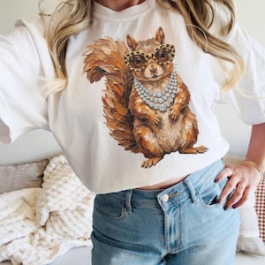 May include: A white t-shirt featuring a central graphic of a brown squirrel with textured brushstroke details. The squirrel is wearing large, round, leopard-print glasses and a multi-layered silver-toned necklace with clear, sparkling stones.