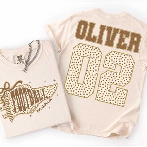 Könnte beinhalten: Zwei cremefarbene T-Shirts. Eines zeigt eine braune Wimpelgrafik mit dem Text "FOOTBALL mama" und Punkten. Das andere zeigt den Namen "OLIVER" in braunen Blockbuchstaben über der großen Zahl "02", die mit braunen Punkten gefüllt ist.